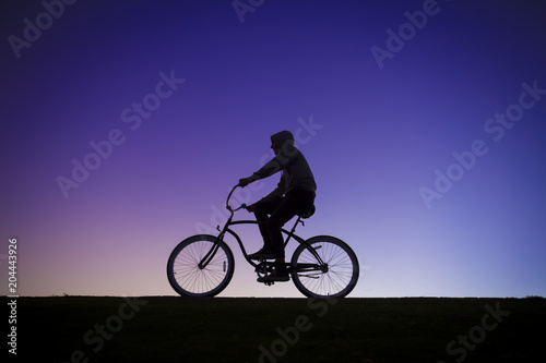 Silhouette of a man in a hoodie riding a cruiser bike in front of a purple sunset sky
