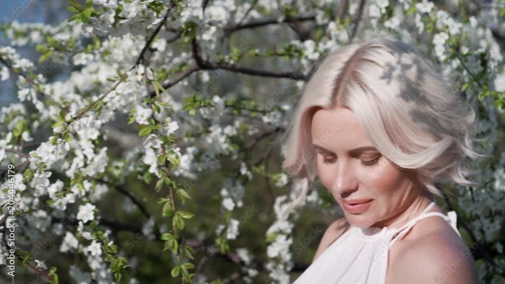Adorable young blonde girl with make-up is lovely smiling and smelling the white flower in the blooming garden. Moving in the wind hair.