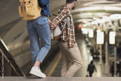 cropped shot of couple of stylish tourists with backpacks holding hands at subway station