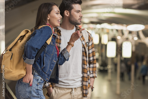 smiling female traveler with backpack pointing by finger to boyfriend at subway station