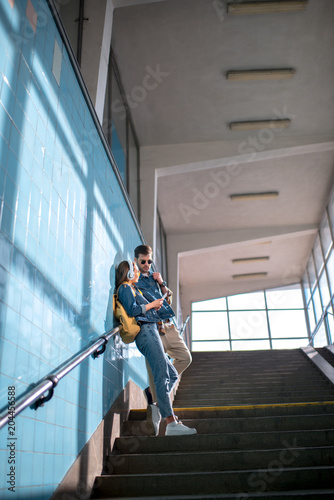 low angle view of stylish female tourist with headphones showing smartphone to boyfriend in sunglasses at subway