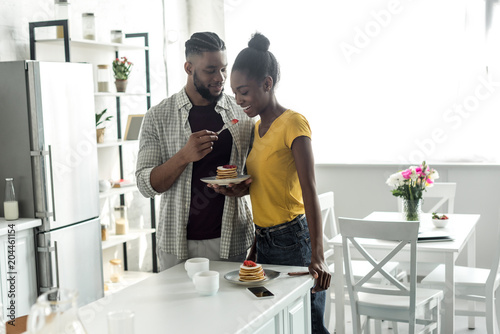 african american boyfriend feeding girlfriend with strawberry at kitchen