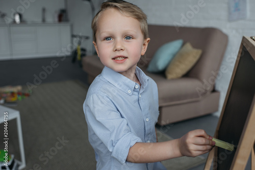 side view of little boy with piece of chalk looking at camera while drawing picture on blackboard at home