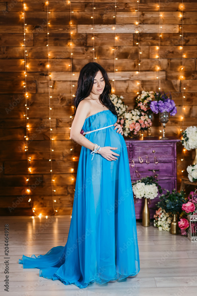 Beautiful pregnant brunette woman in a long silk blue dress standing at full height and looks at the belly