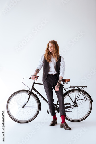 stylish tattooed businessman with curly hair leaning on bicycle and looking away