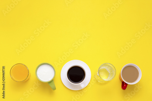 Group of Drinks in Multicolored Cups, Black Coffee, Coffee with Milk, Yogurt, Just Water, Orange Juice on Yellow Background. Flat lay, top view