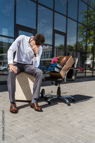 young depressed manager with box of personal stuff sitting on chair outdoors after he was fired