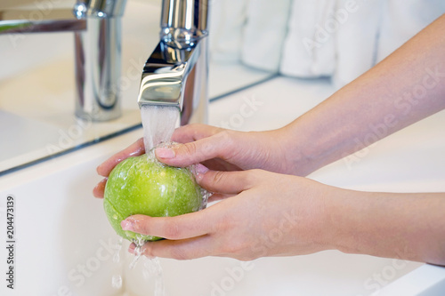 Female hands wash the apple under the tap. Woman young housewife washing fresh green apple in kitchen under water stream. Healthy eating.