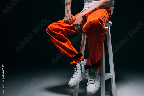 Cropped view of man in prison uniform with cuffs on legs sitting on stool on dark background