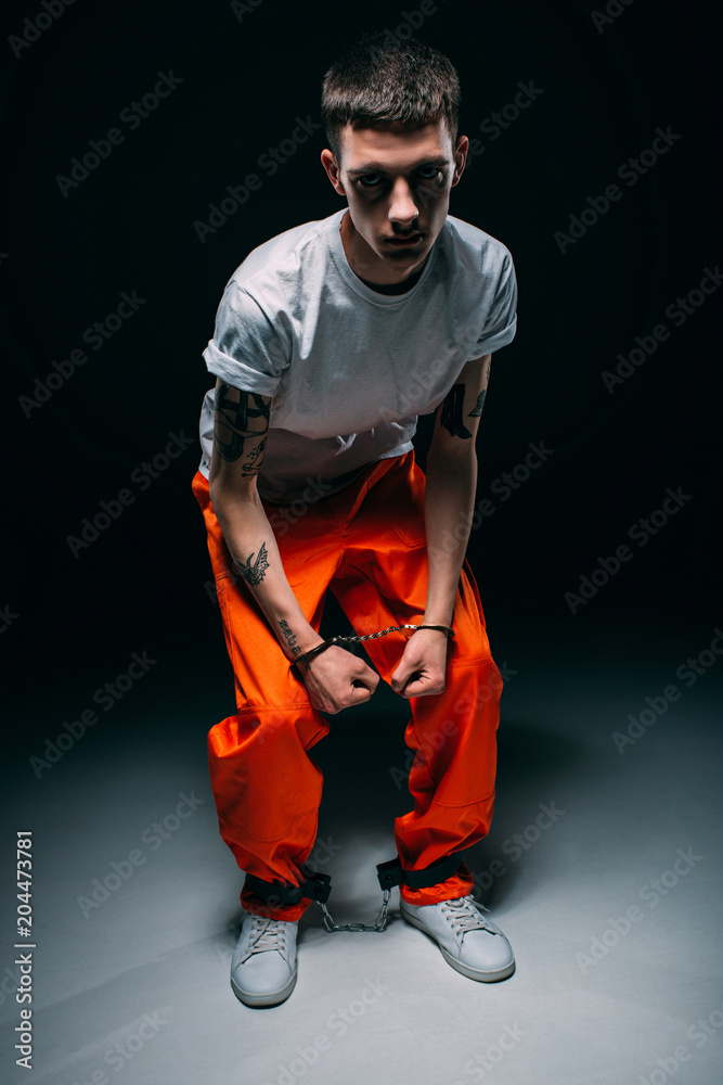 Sad male prisoner in orange uniform and cuffs on dark background Sad male prisoner in orange uniform and cuffs on dark background