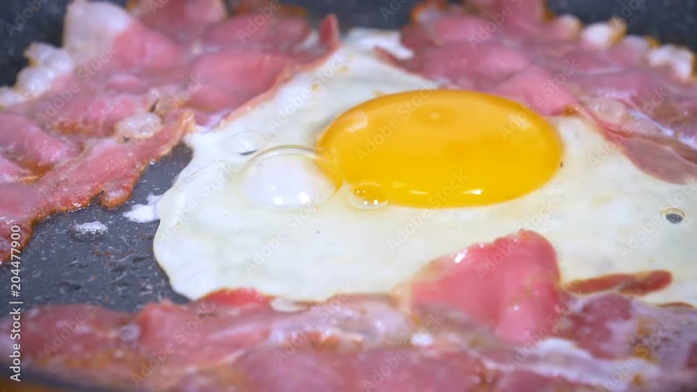 Egg being dropped on fried bacon for breakfast in slow motion. Egg being dropped on fried bacon for breakfast in slow motion.