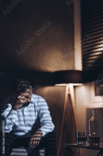 Silhouette of a stressed middle-aged man with problems sitting alone with his head down next to a bottle and a glass of alcohol in a dark room with window blinds and a dimmed light of a standing lamp