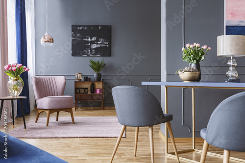 Grey chair at table and pink armchair in open space interior with black painting and flowers