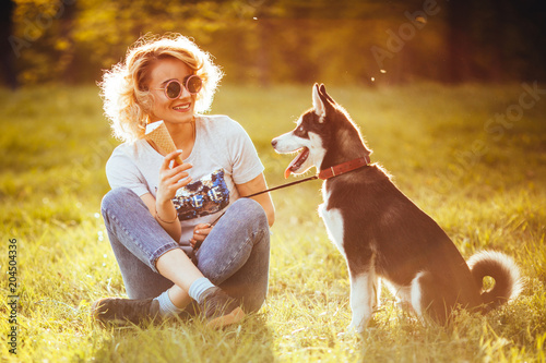 a girl in jeans and in white T-shirt with ice cream in her hand sits cross-legged in the meadow and looks at a pet in the park in the summer