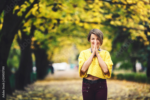 Fototapeta Naklejka Na Ścianę i Meble -  beautiful girl in autumn stands on alley with yellow maples and holds her hands near her mouth with smile.