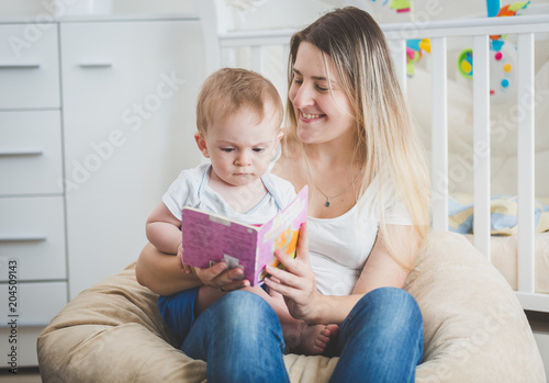 Beautiful smiling young mother reading fairy tale to her baby boy sitting on her lap