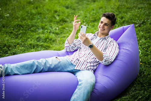 Handsome young man lying on inflatable sofa lamzac use on phone while resting on grass in park photo