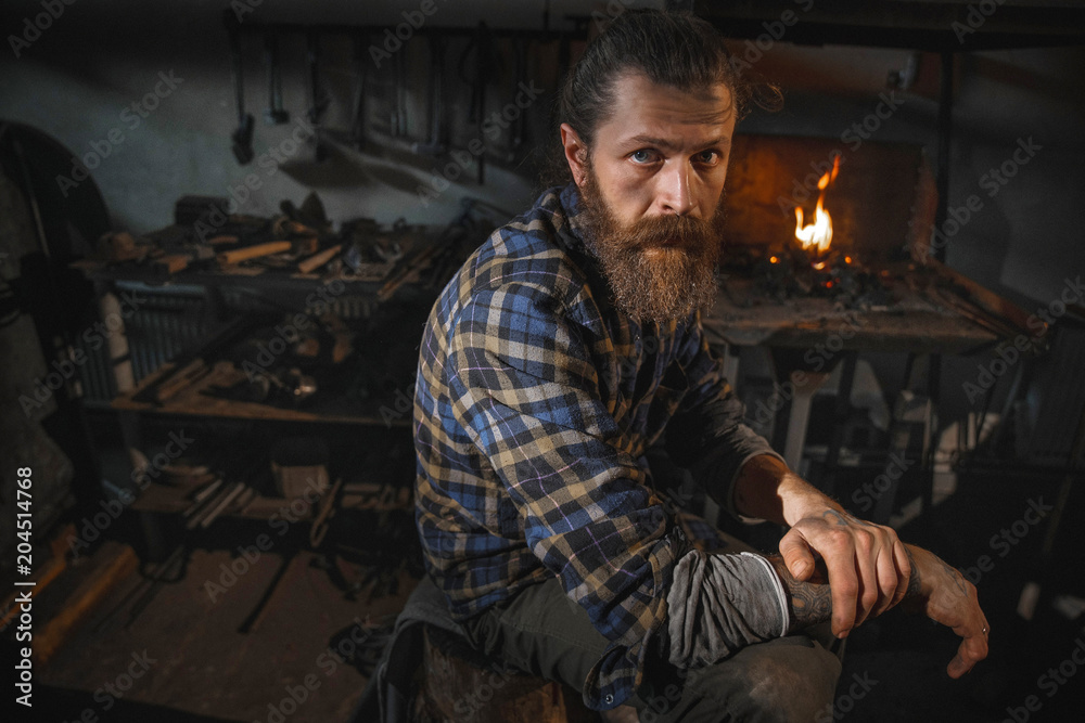 Portrait of a real Brutal blacksmith of a man with a beard after working in his workshop against the background of a burning flame. Portrait of a profession.