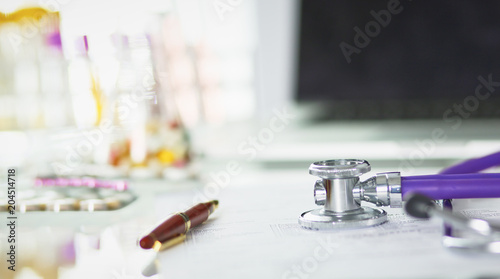 closeup of the desk of a doctors office with a stethoscope in the foreground and a bottle with pills in the background, selective focus