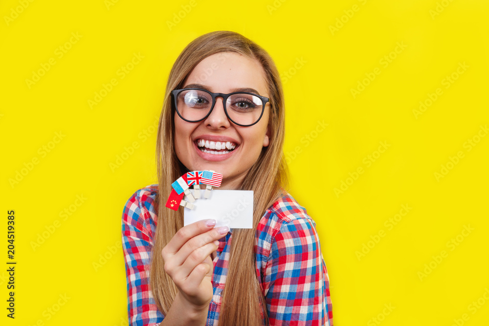 Young woman with card and flags of different speaking countries. Studio portrait of young beautiful female student on yellow background Young woman with card and flags of different speaking countries. Studio portrait of young beautiful female student on yellow background