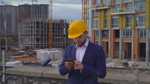 young attractive man in a yellow helmet makes photos of buildings under construction on a mobile phone. the person sends messages to the customer via a smartphone. 4k