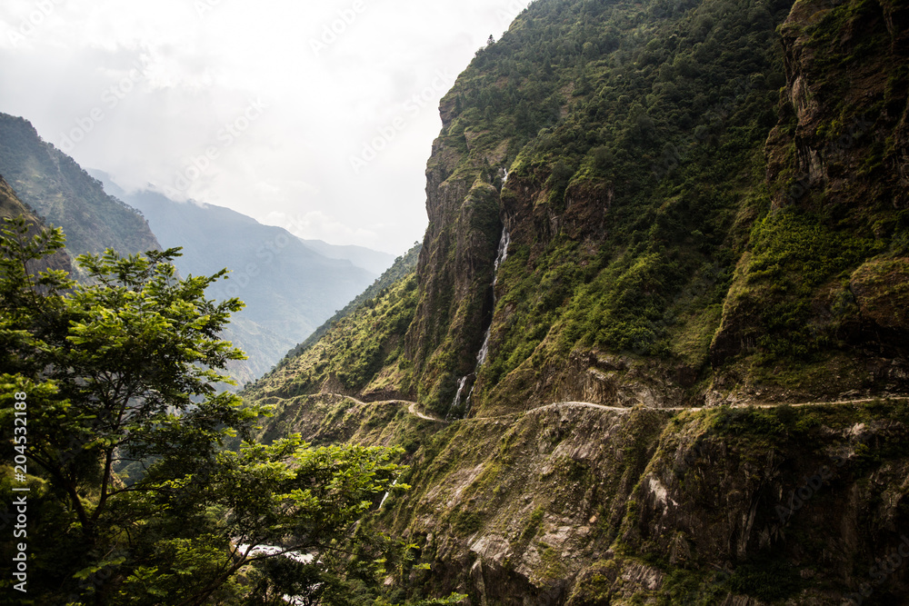 Scenic Himalayan Landscape. View of Waterfall, Mountains and the road with forest and green trees. Annapurna Range on Annapurna Circuit Trek. Autumn season in Nepal, Asia.
