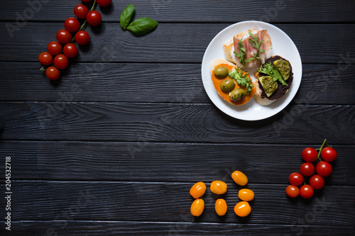 Tasty bruschettas with parma, rucola, olive pasta, olives, humus and basil on white plate on black wooden background. Italian cuisine. Cherry red and orange tomatoes. Copy space, mock up. Top view.