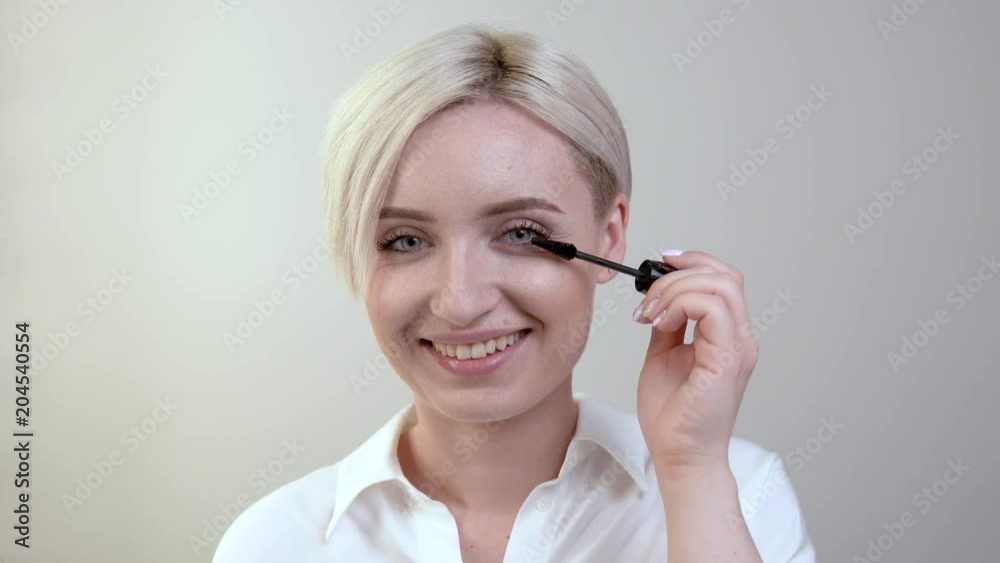 Cute shorthaired blonde girl looks directly at the camera. Applies mascara on top of lashes of left eye. Cute shorthaired blonde girl looks directly at the camera. Applies mascara on top of lashes of left eye.