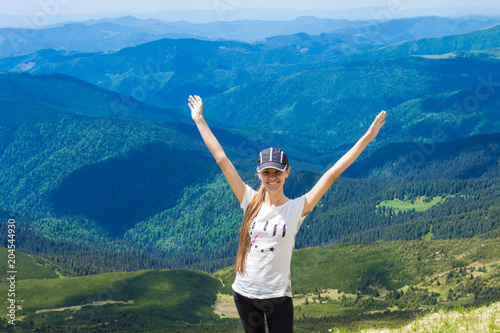 Happy young woman with hands up stands on top of hill  against backdrop of beautiful mountains and blue sky on summer day. Holiday travel concept