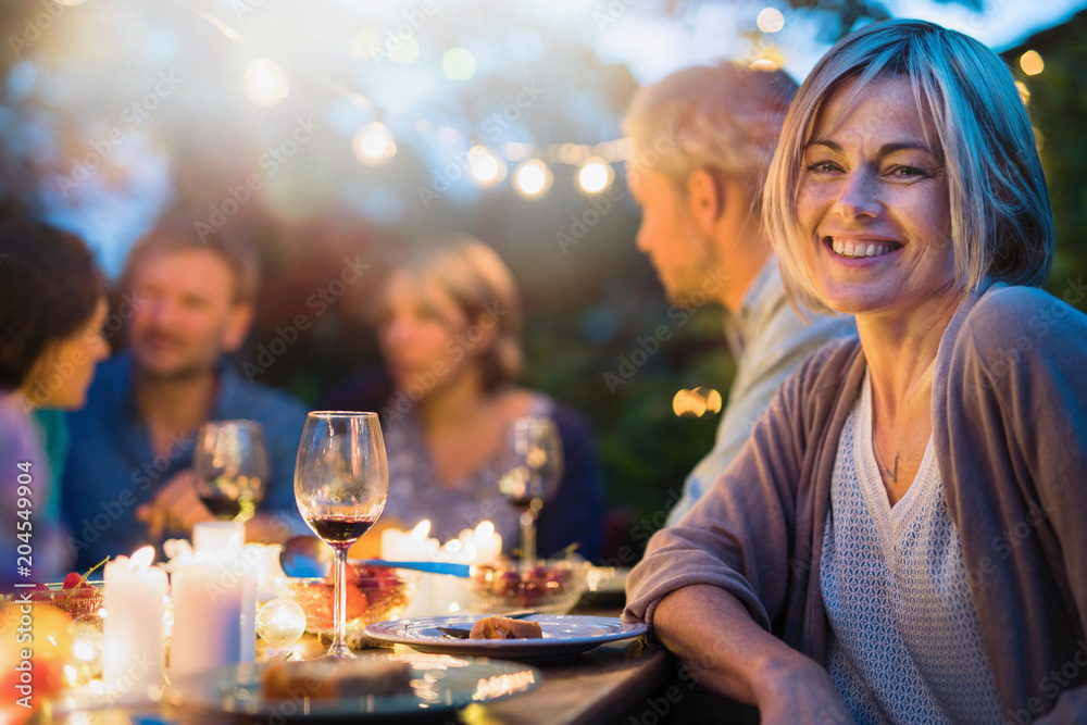 Group of friends gathered around a table in a garden on a summer evening to share a meal and have a good time together. Focus on a beautiful woman - obrazy, fototapety, plakaty 