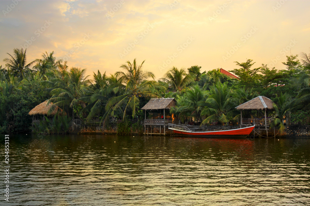 Pristine riverbank at sunset, palms trees, a red boat and bungalows, situated at the Praek Tuek Chhu River in Kampot Province in southern Cambodia, Asia. Natural asian riverside landscape
