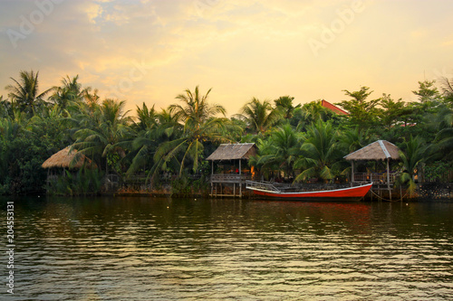Pristine riverbank at sunset, palms trees, a red boat and bungalows, situated at the Praek Tuek Chhu River in Kampot Province in southern Cambodia, Asia. Natural asian riverside landscape © Daniel