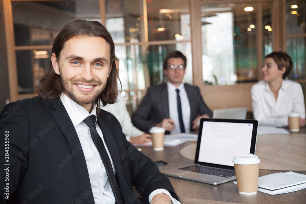 Smiling handsome young businessman in suit and tie looking at camera posing at group meeting with partners, happy executive manager, successful team leader or professional business coach portrait