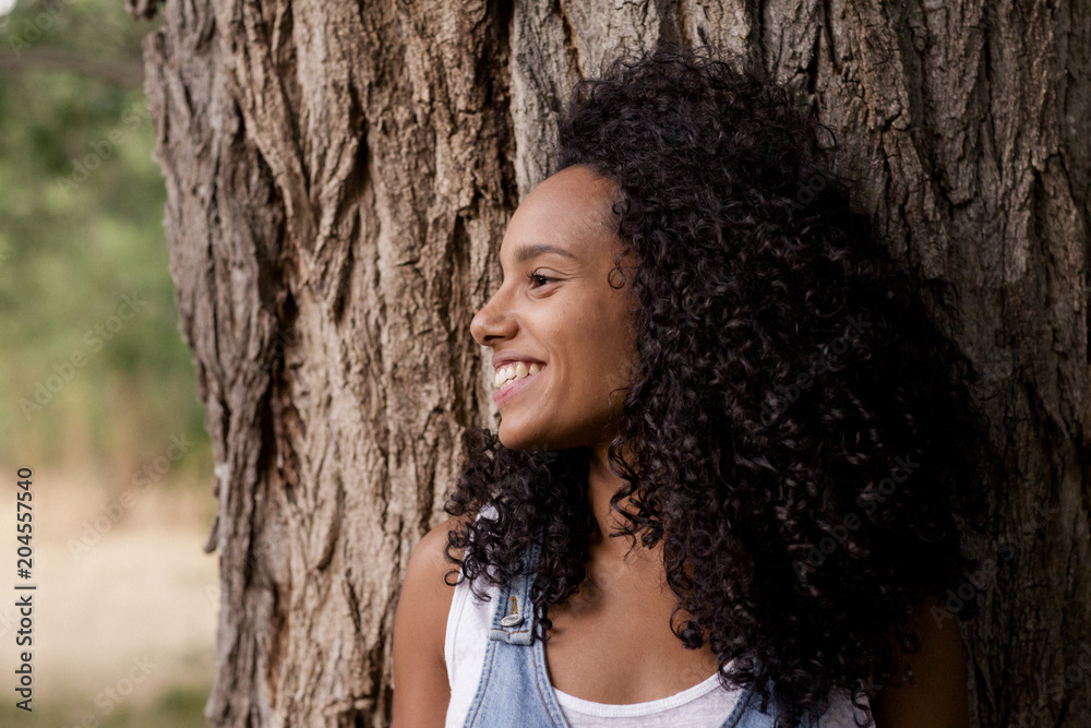 portrait outdoors of a beautiful young afro american woman smiling at sunset. Brown tree background. Lifestyle in nature. Casual jean dress clothing portrait outdoors of a beautiful young afro american woman smiling at sunset. Brown tree background. Lifestyle in nature. Casual jean dress clothing
