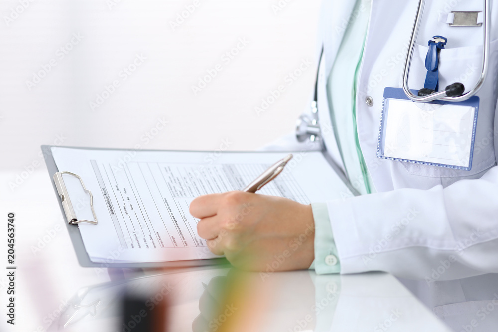 Female doctor filling up an application form with silver pen while sitting at the desk in hospital office. Just hands. Medicine and health care concept