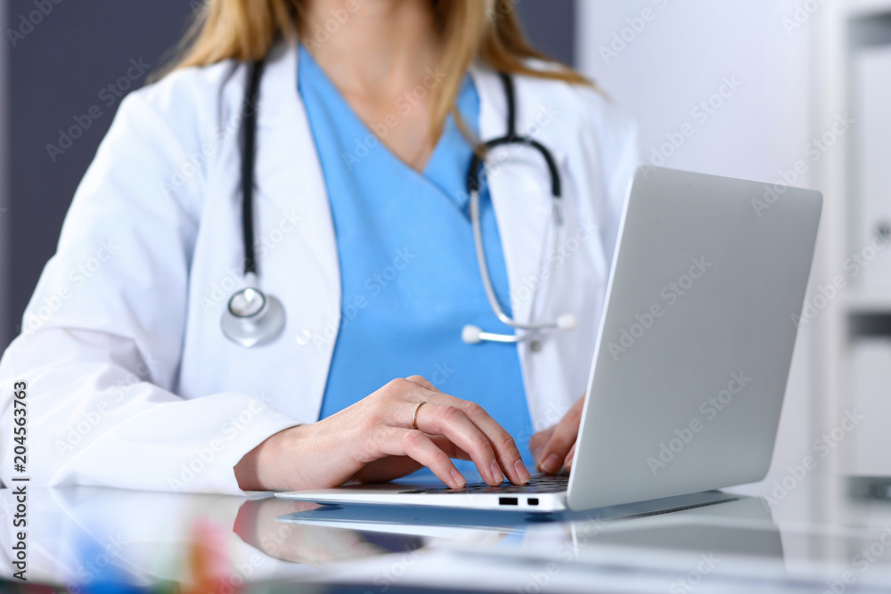 Doctor typing on laptop computer while sitting at the glass desk in hospital office. Physician at work. Medicine and healthcare concept