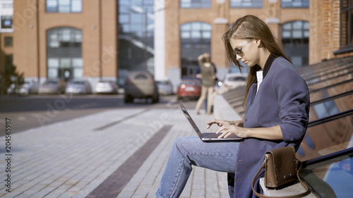 A young pretty concentrated girl in jeans in sunglasses is working with a laptop outdoors near a loft