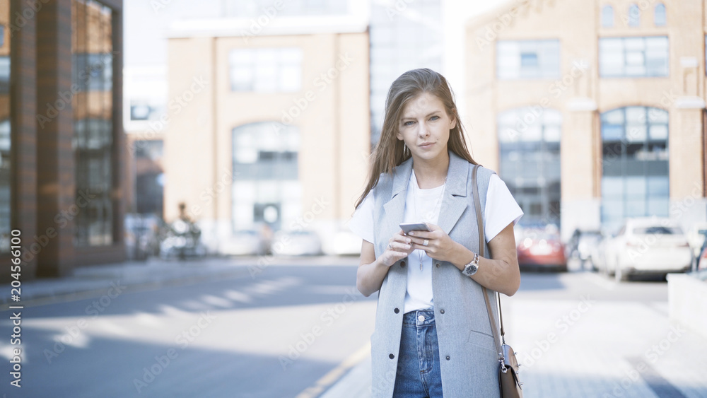 A young girl alone dressed in a jacket holding a phone looking at the camera outdoors