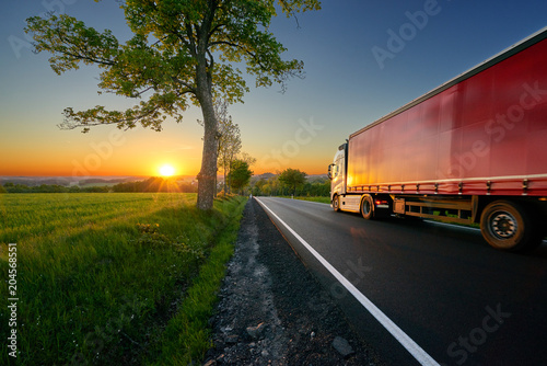 Truck driving on the asphalt road between trees in a rural landscape at sunset