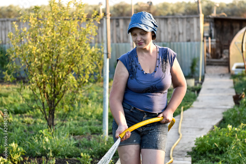 Sensual caucasian woman pours water from a hose flowers and vegetables, trees in her garden. A hot summer day, a river in the background, a greenhouse, an excellent harvest of fruits and vegetation. photo