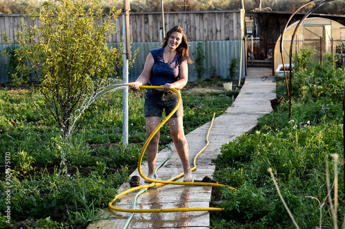 Sensual caucasian woman pours water from a hose flowers and vegetables, trees in her garden. A hot summer day, a river in the background, a greenhouse, an excellent harvest of fruits and vegetation. photo