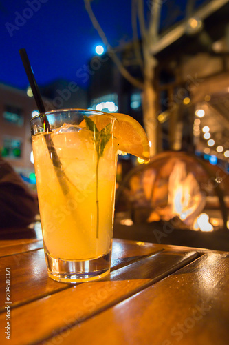 Refreshing cocktail beverage with ice and straw scene from outdoor restaurant at night with lights and firepit. 