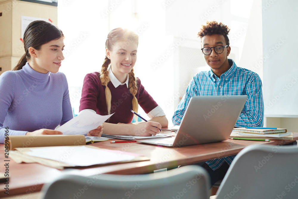 Three friendly students sitting by desk, looking at laptop display and reading online information while preparing seminar tasks Three friendly students sitting by desk, looking at laptop display and reading online information while preparing seminar tasks