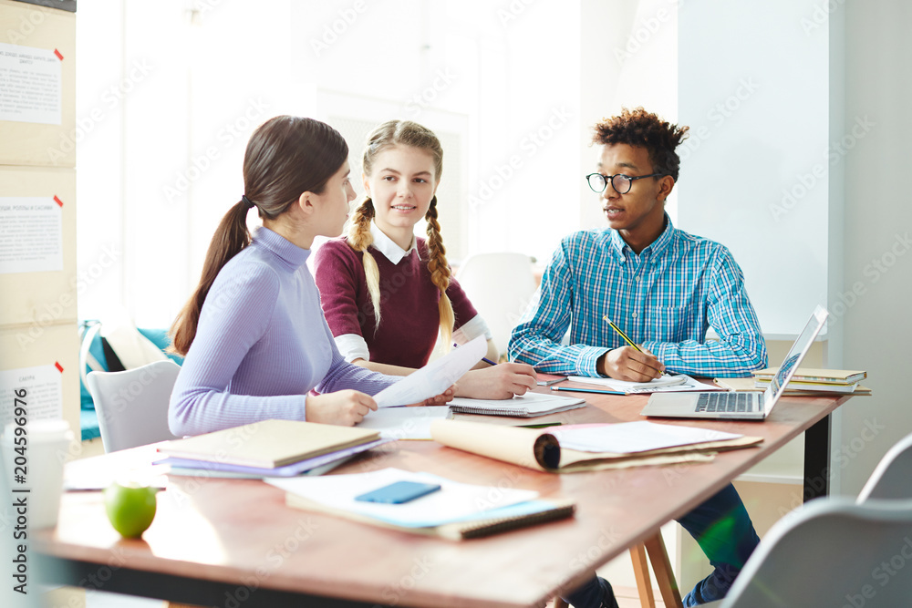 Team of modern students of college or high school having discussion of home assignment while preparing it Team of modern students of college or high school having discussion of home assignment while preparing it