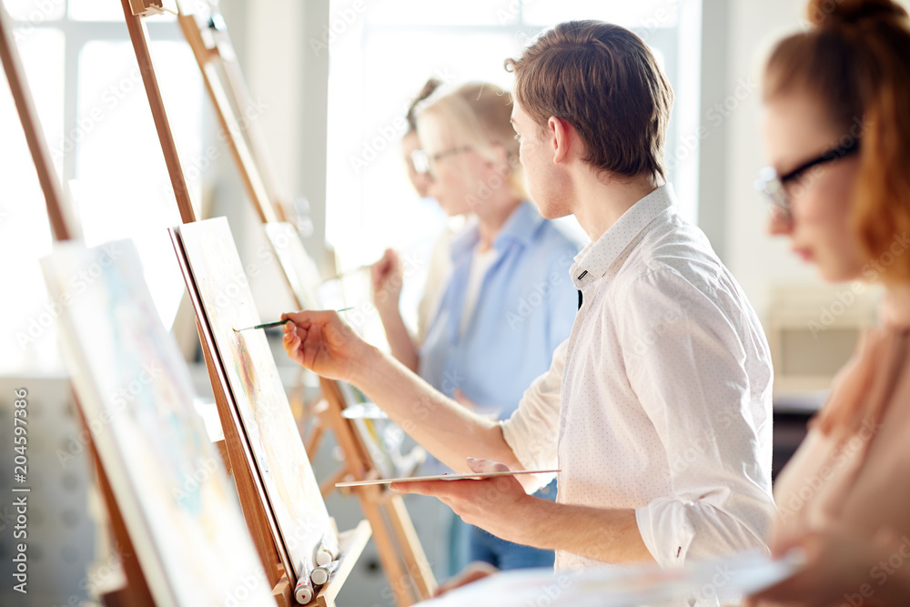 Creative guy looking at picture on easel of teacher while painting at lesson in studio of arts Creative guy looking at picture on easel of teacher while painting at lesson in studio of arts