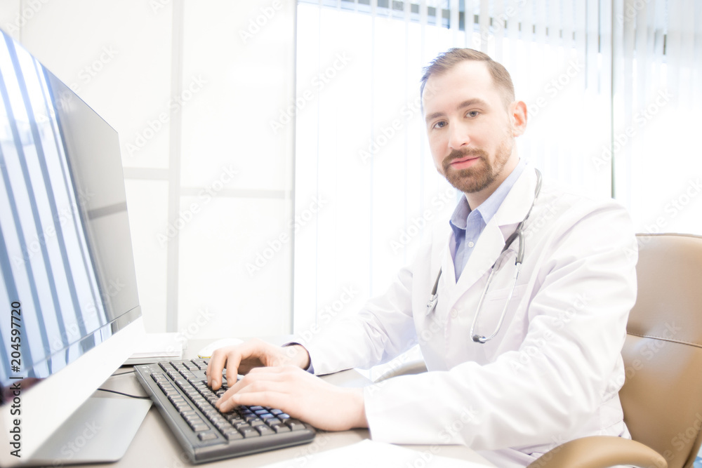 Young doctor looking at camera while sitting by computer monitor during online consultation