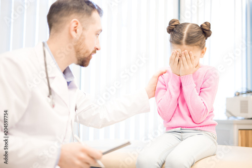 Male clinician comforting crying little girl during medical appointment in hospital