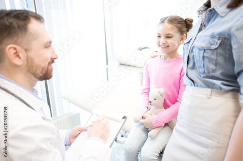 Happy girl with teddybear and her mother talking to doctor while visiting modern clinics