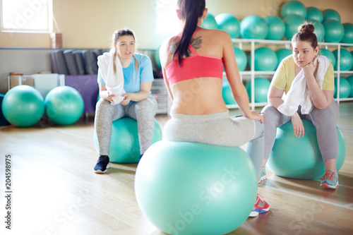 Back view of young instructor sitting on fitball in front of two chubby girls consulting about workout