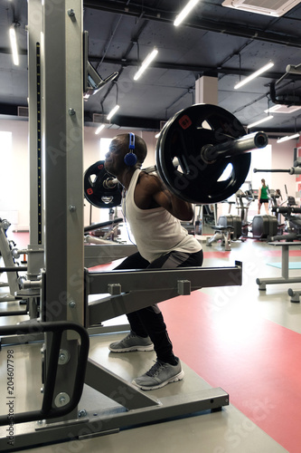 A black muscular guy in a white tank top, in blue headphones squats with a barbell.